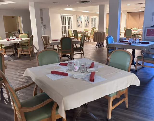 A dining room with multiple tables covered in white tablecloths, each set with plates, glasses, and red cloth napkins. The room has wooden flooring, green cushioned chairs with wooden frames, and soft lighting. There are paintings on the far wall and a sign visible in the background.