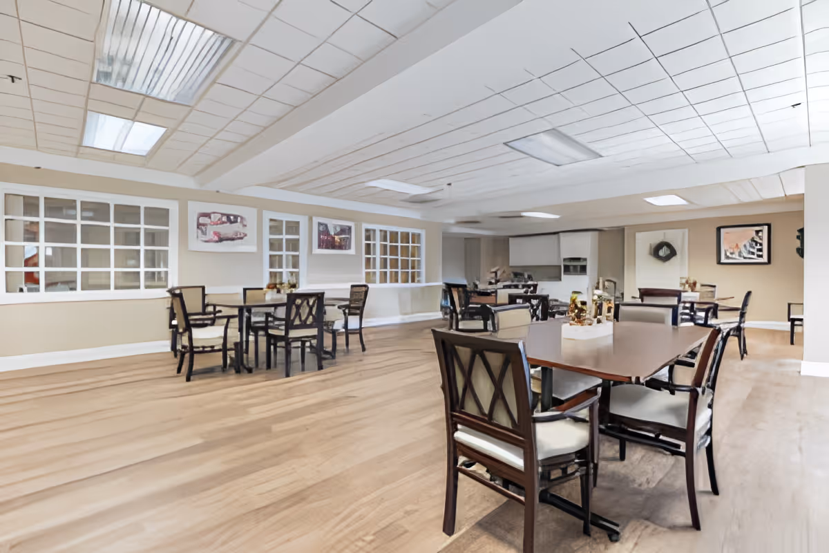 A spacious dining room with multiple wooden tables and cushioned chairs arranged neatly. The room has light-colored walls, large windows with white frames, and several framed pictures on the walls. The ceiling has recessed lighting panels, and the floor is covered with wood-like flooring.