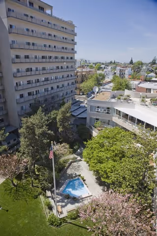 View of a multi-story residential building surrounded by trees and greenery, with a small pool and an American flag in the courtyard below. Other buildings and houses are visible in the background under a clear blue sky.