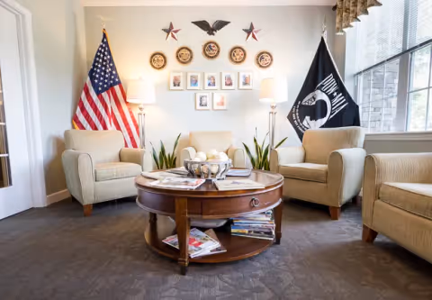 A cozy sitting area with four beige armchairs arranged around a round wooden coffee table filled with magazines. The wall behind features two flags, an American flag on the left and a POW/MIA flag on the right, along with decorative stars, circular plaques, and framed photos. Two floor lamps provide soft lighting, and a large window on the right lets in natural light.