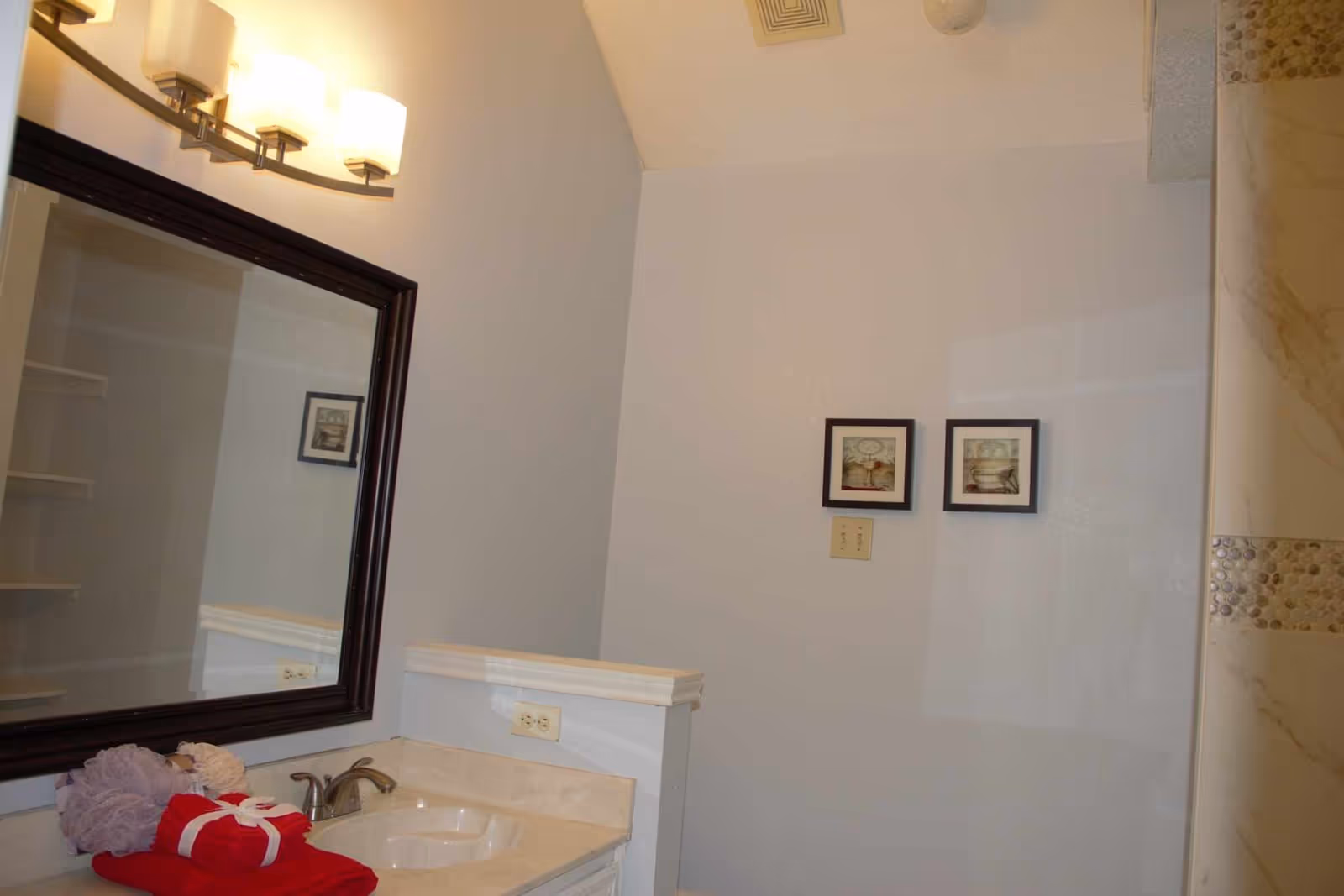 Bathroom vanity with sink, mirror and wall light, folded towels on the counter and framed pictures beside a tiled shower.