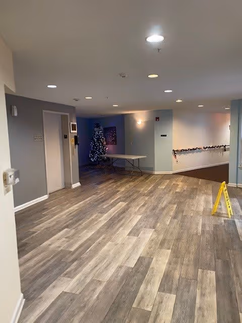 Interior common-area hallway with wood-look flooring, recessed ceiling lights, a folding table, a decorated Christmas tree in the back, and a yellow caution sign.