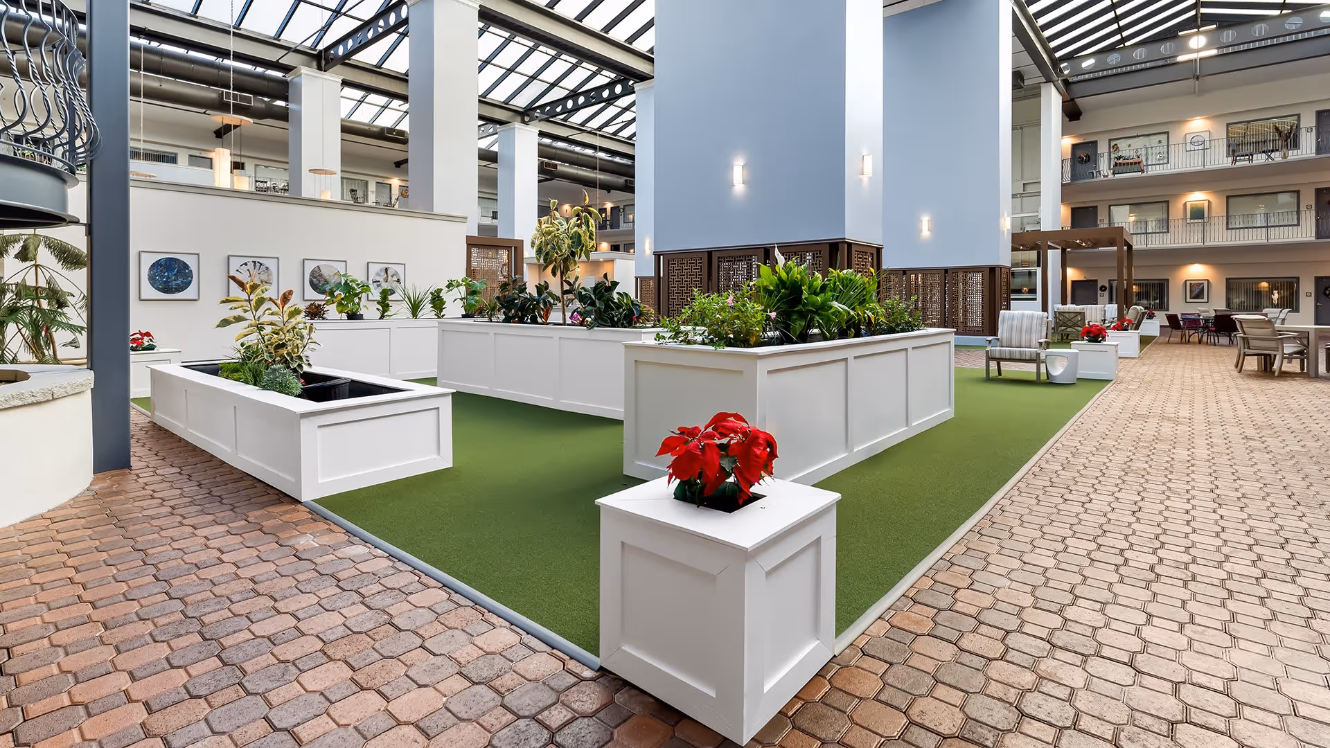 Indoor atrium area with raised white planter boxes filled with various green plants and red flowers. The space has a green carpeted section surrounded by a brick-patterned floor. There are seating areas with chairs and tables along the right side, and multiple floors of rooms with balconies overlooking the atrium. The ceiling is glass, allowing natural light to illuminate the area.