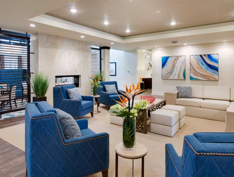 Bright, modern lounge area with blue upholstered armchairs, a beige sofa, coffee tables, a vase of flowers and wall art.