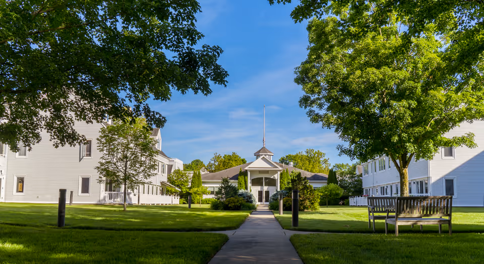 A sunny outdoor view of Pomperaug Woods senior living facility with a paved walkway leading to a central building surrounded by green lawns, trees, and benches. Two white multi-story buildings flank the walkway on either side under a clear blue sky.
