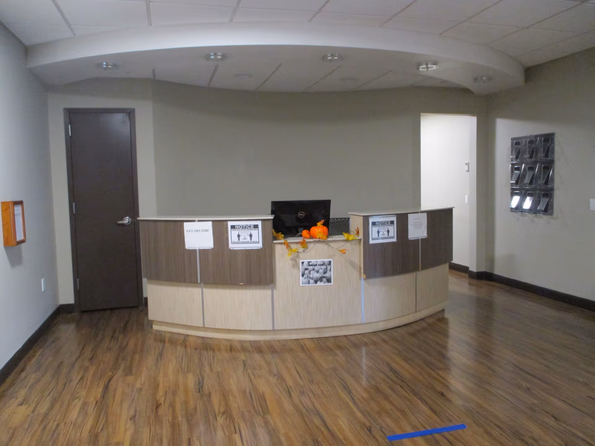 Reception desk area with a computer monitor, autumn-themed decorations including small pumpkins and leaves, and several notices posted on the front. The room has wood flooring, beige walls, a closed brown door to the left, and a wall-mounted brochure holder on the right.