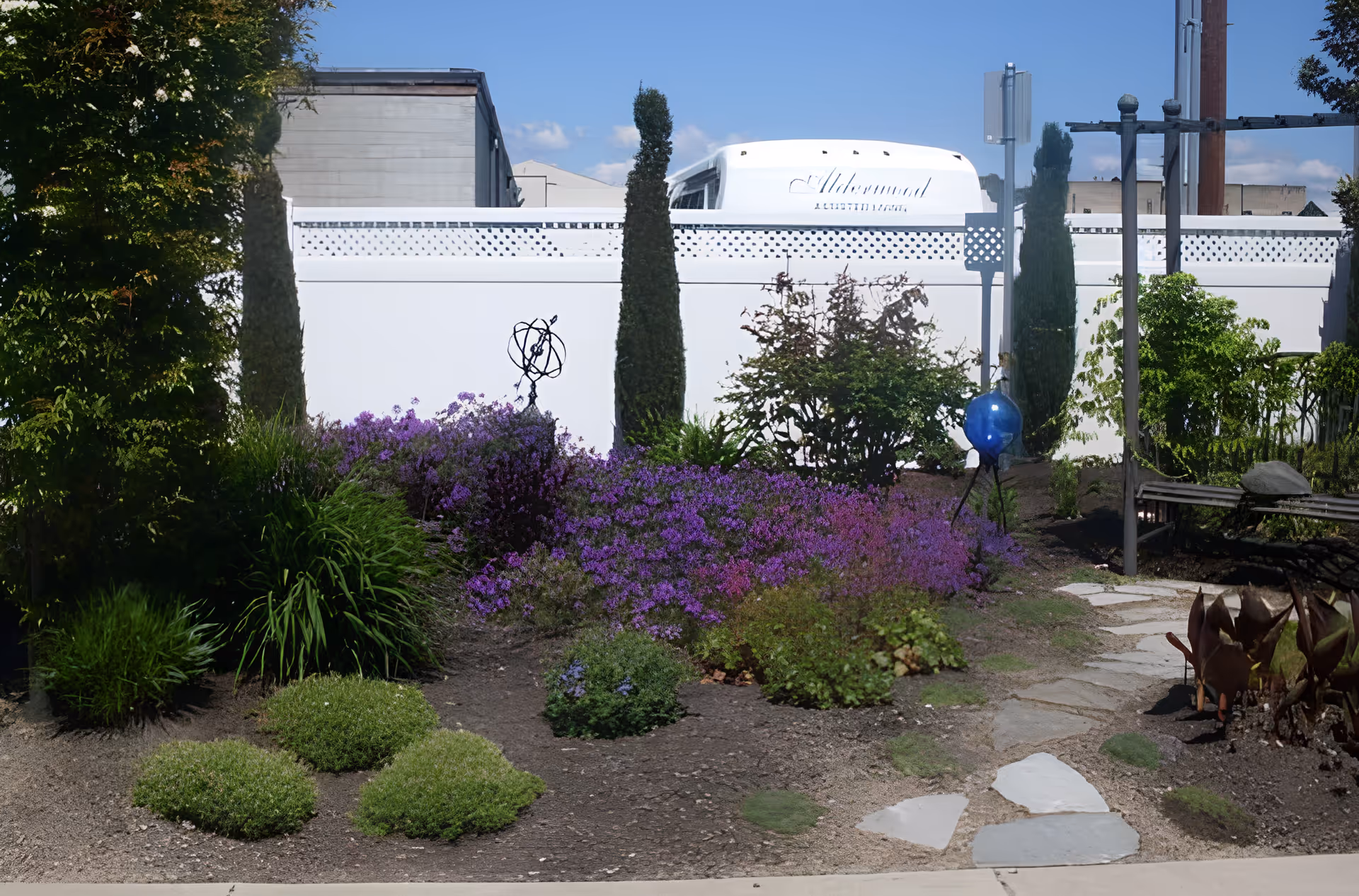 A garden area with various green plants and purple flowers, a stone pathway, and a white fence in the background. Behind the fence, the top of a white vehicle with the text 'Alderwood Assisted Living' is visible. The sky is clear and blue.
