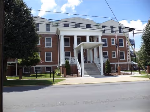 Front exterior view of a three-story brick nursing home building with white columns at the entrance, a covered stairway, a fenced lawn, and an American flag on a flagpole to the right.