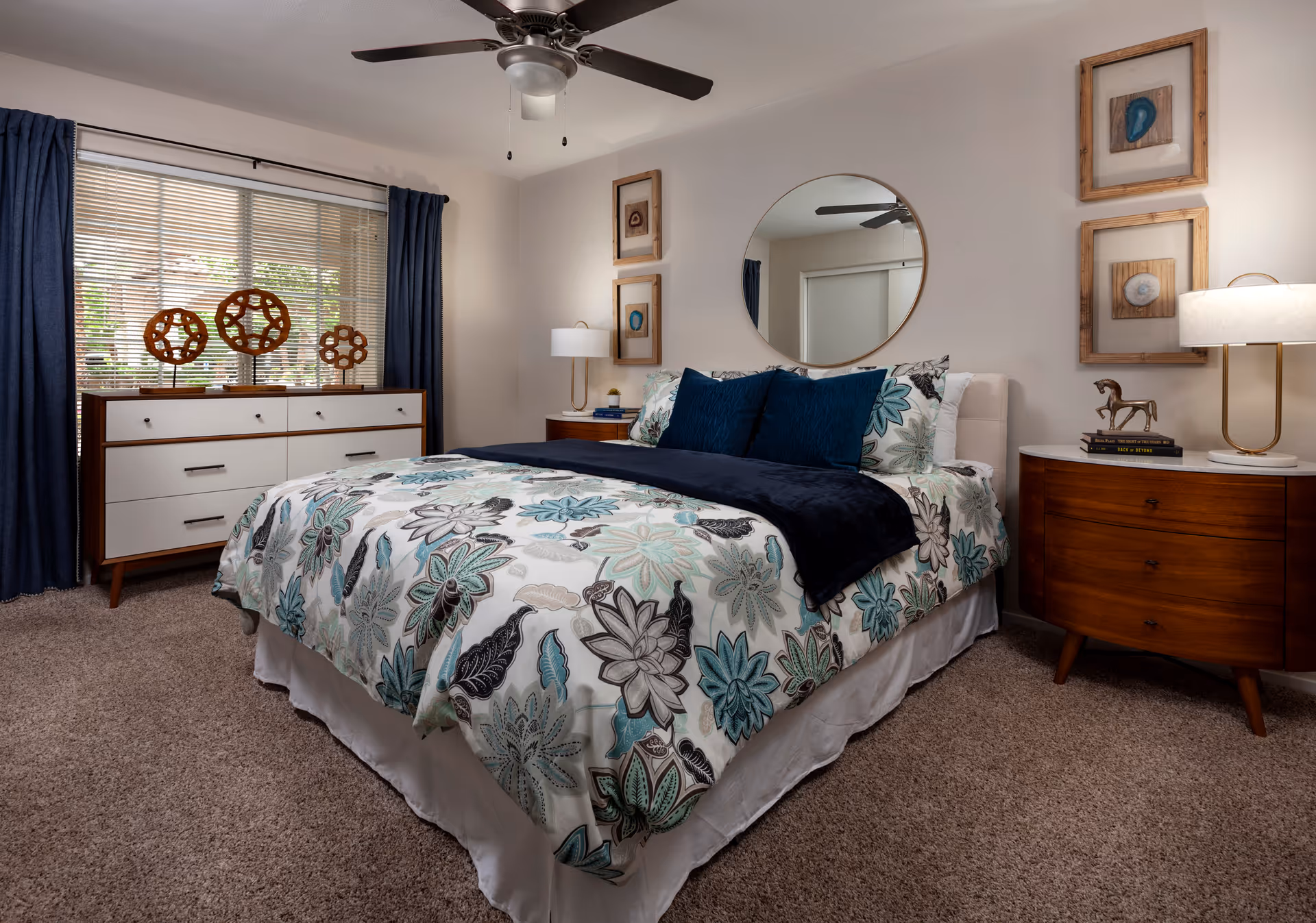 A neatly made bed with floral patterned bedding and dark blue accent pillows in a bedroom. The room features a round mirror above the bed, two wooden nightstands with lamps, framed artwork on the walls, a dresser with decorative wooden sculptures, and a window with blinds and dark blue curtains. The floor is carpeted and there is a ceiling fan with a light fixture.