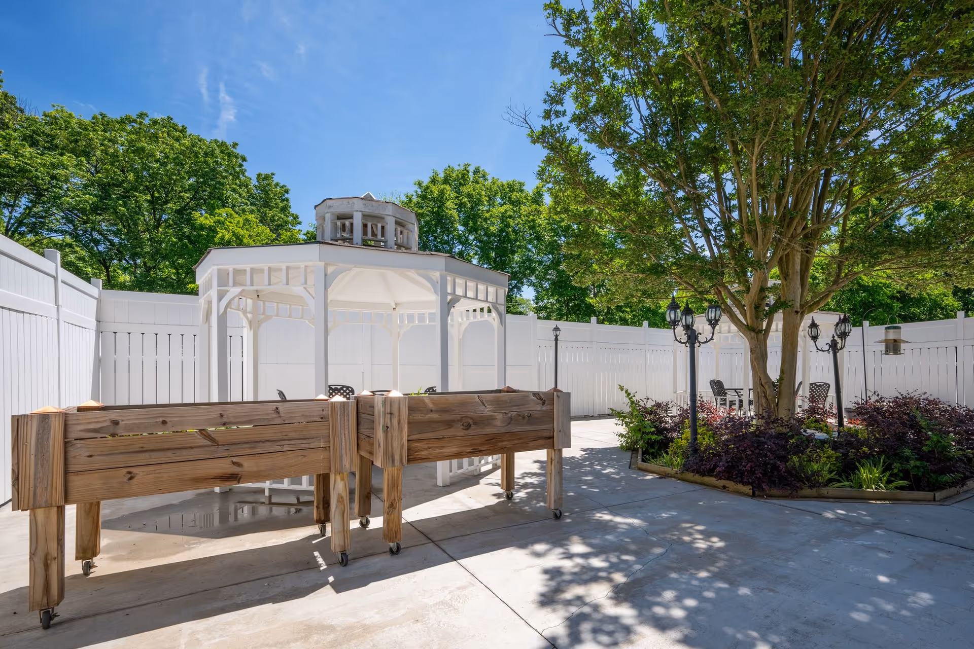 Outdoor courtyard area with a white gazebo, wooden raised garden beds on wheels, a large tree surrounded by plants, black vintage-style lamp posts, and a white privacy fence under a clear blue sky.
