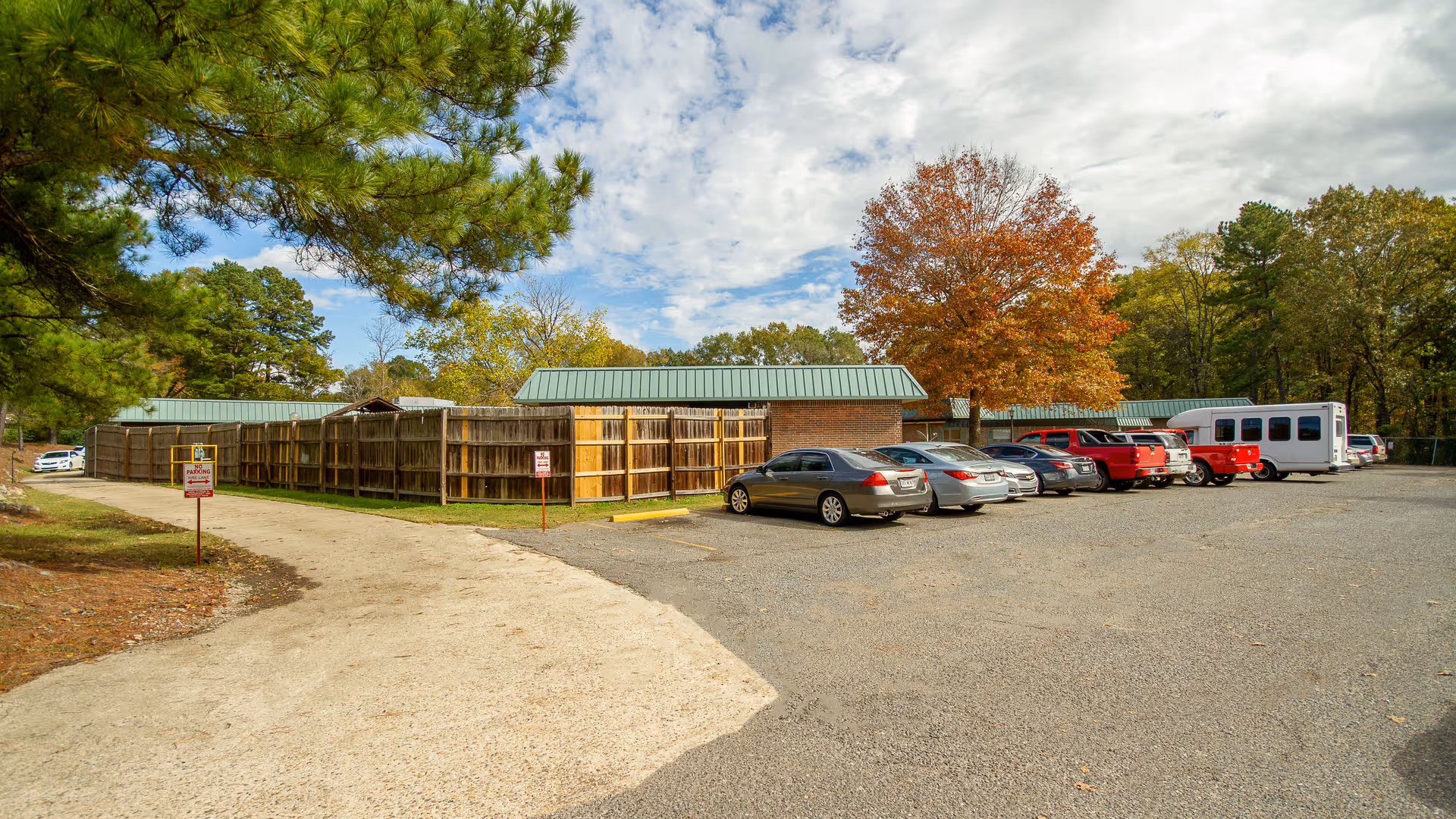 Parking lot outside a single-story brick building with green metal roofs, surrounded by trees with autumn foliage. Several cars and a white shuttle bus are parked in the lot. A wooden fence encloses part of the area, and the sky is partly cloudy.