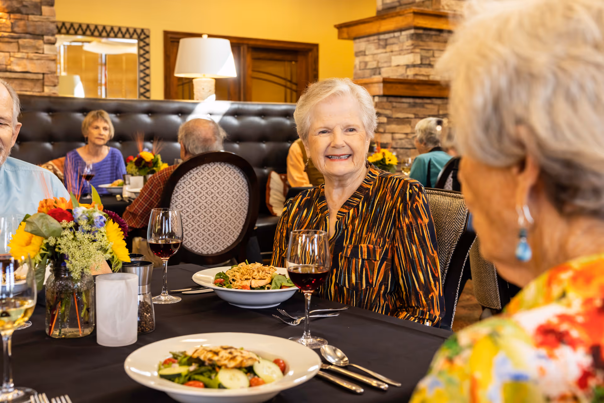 A group of elderly people dining together in a warmly decorated restaurant or dining room. The focus is on a smiling elderly woman wearing a patterned blouse, sitting at a table with plates of salad and glasses of wine. The background shows other diners and cozy interior elements like a stone fireplace and a lamp.
