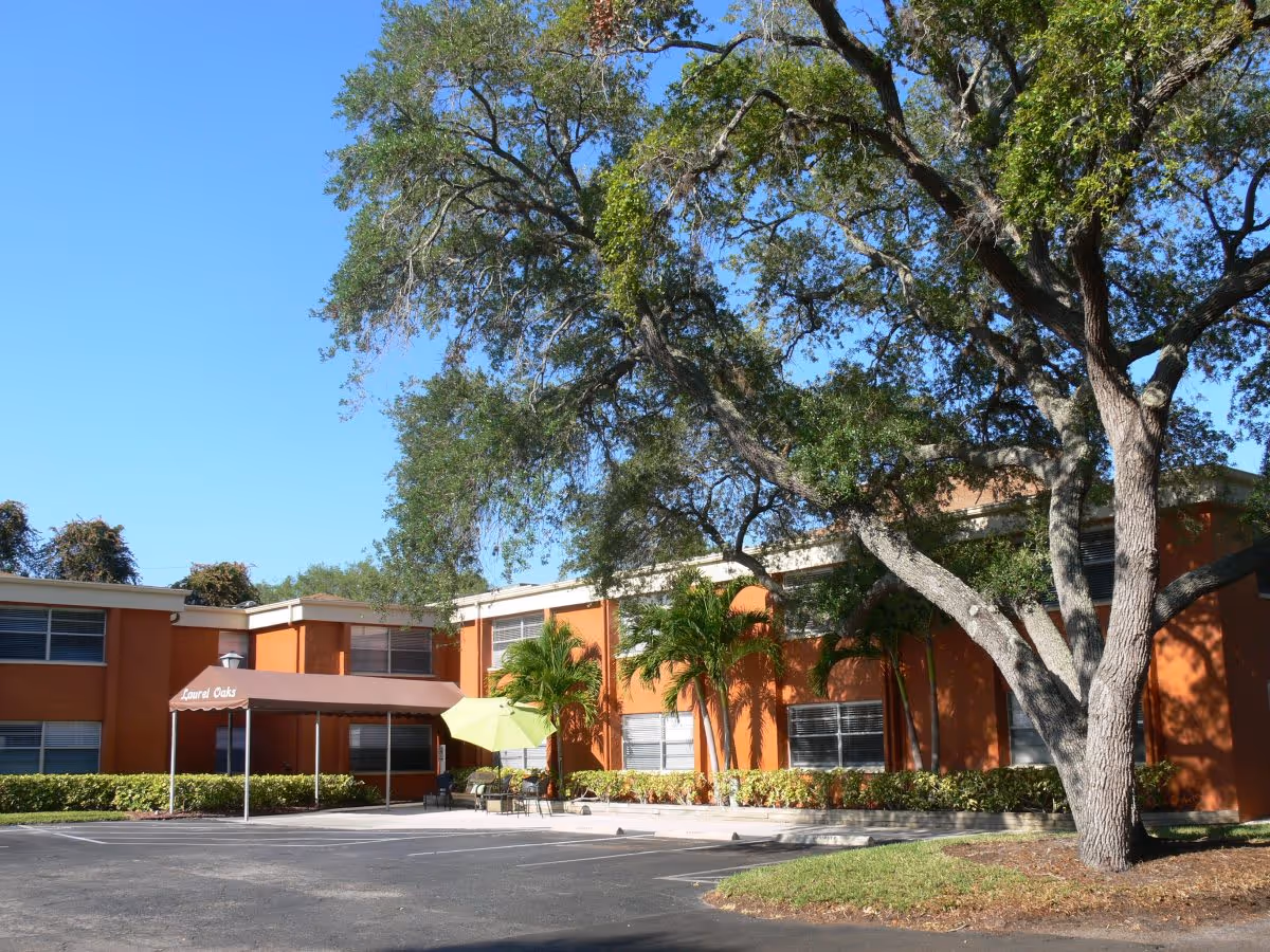Front exterior of an orange two-story senior living building with a covered entrance, patio seating, a large tree, and a parking lot.
