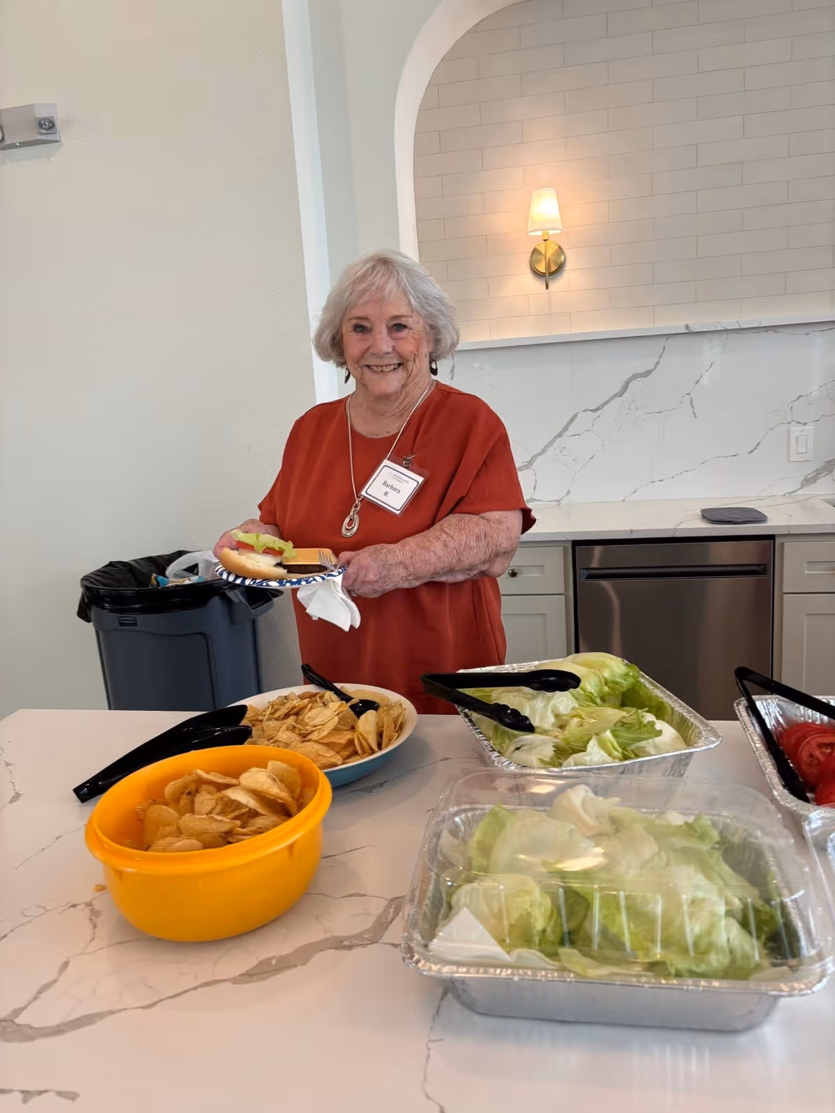 An elderly woman with gray hair wearing a red shirt and a name tag stands behind a kitchen counter with bowls of potato chips, lettuce, and sliced tomatoes. She is holding a plate with a sandwich and smiling. The kitchen has white marble countertops and a modern backsplash with a wall sconce light.