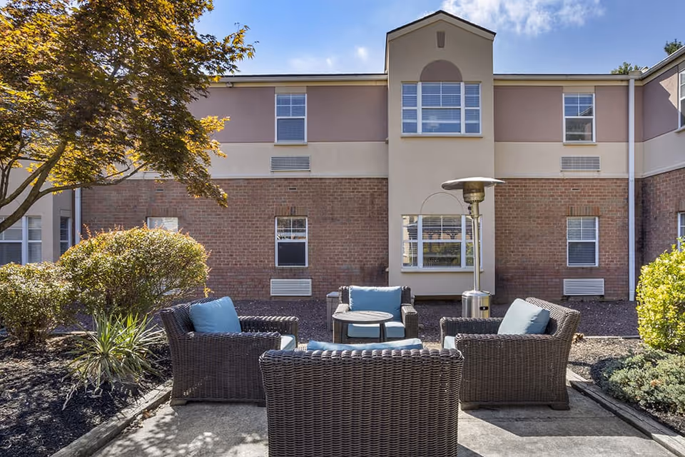 Outdoor patio area with four wicker chairs with blue cushions arranged around a small round table. There is a tall outdoor heater next to the seating area. The patio is surrounded by bushes and trees, with a two-story brick and beige building in the background under a clear blue sky.