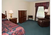 A bedroom in Henry County Nursing Home featuring a bed with a patterned bedspread, a wooden nightstand with a lamp, a wooden dresser, a small table, a window with a valance, and a brown recliner chair on a green carpeted floor.