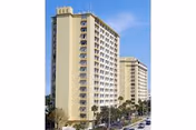 Exterior view of two tall, beige residential buildings with multiple windows under a clear blue sky, surrounded by palm trees and a street with cars.