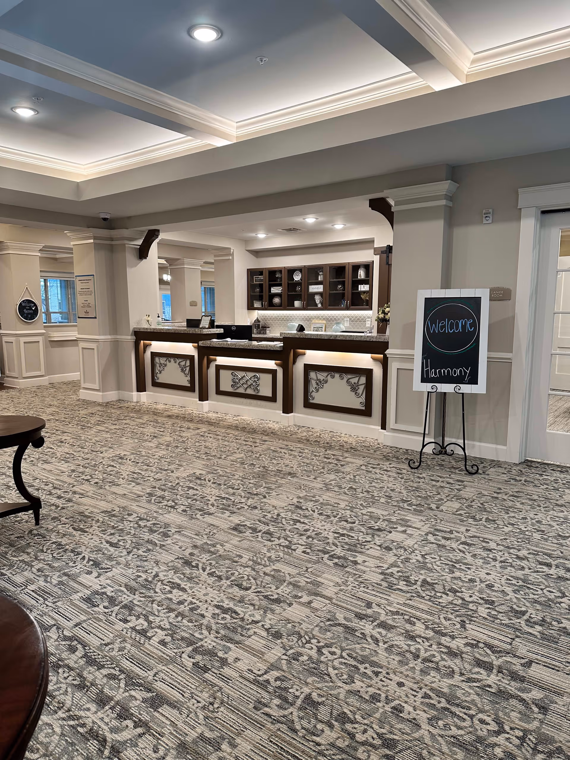 Interior view of a senior living facility reception area with a patterned carpet, a counter with decorative panels, and shelves with decorative items behind the counter. A chalkboard sign on a stand reads 'Welcome Harmony'. The ceiling has recessed lighting and crown molding.