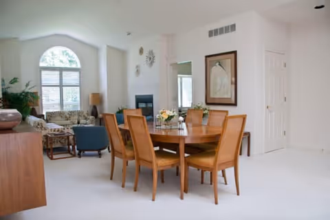 A bright and spacious living and dining area with a wooden dining table and six chairs in the foreground. In the background, there is a seating area with patterned and blue armchairs, a large arched window with blinds, a fireplace, and decorative wall art. The walls are painted white, and the room has a clean, minimalist design.