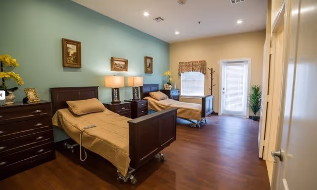A well-lit bedroom with two hospital-style beds, each with beige bedding and wooden headboards and footboards. Between the beds are two nightstands with lamps. The walls are painted in light blue and beige tones, decorated with framed pictures. There is a window with blinds and a door with glass panels leading outside. The room has wooden flooring and some decorative plants and flowers.
