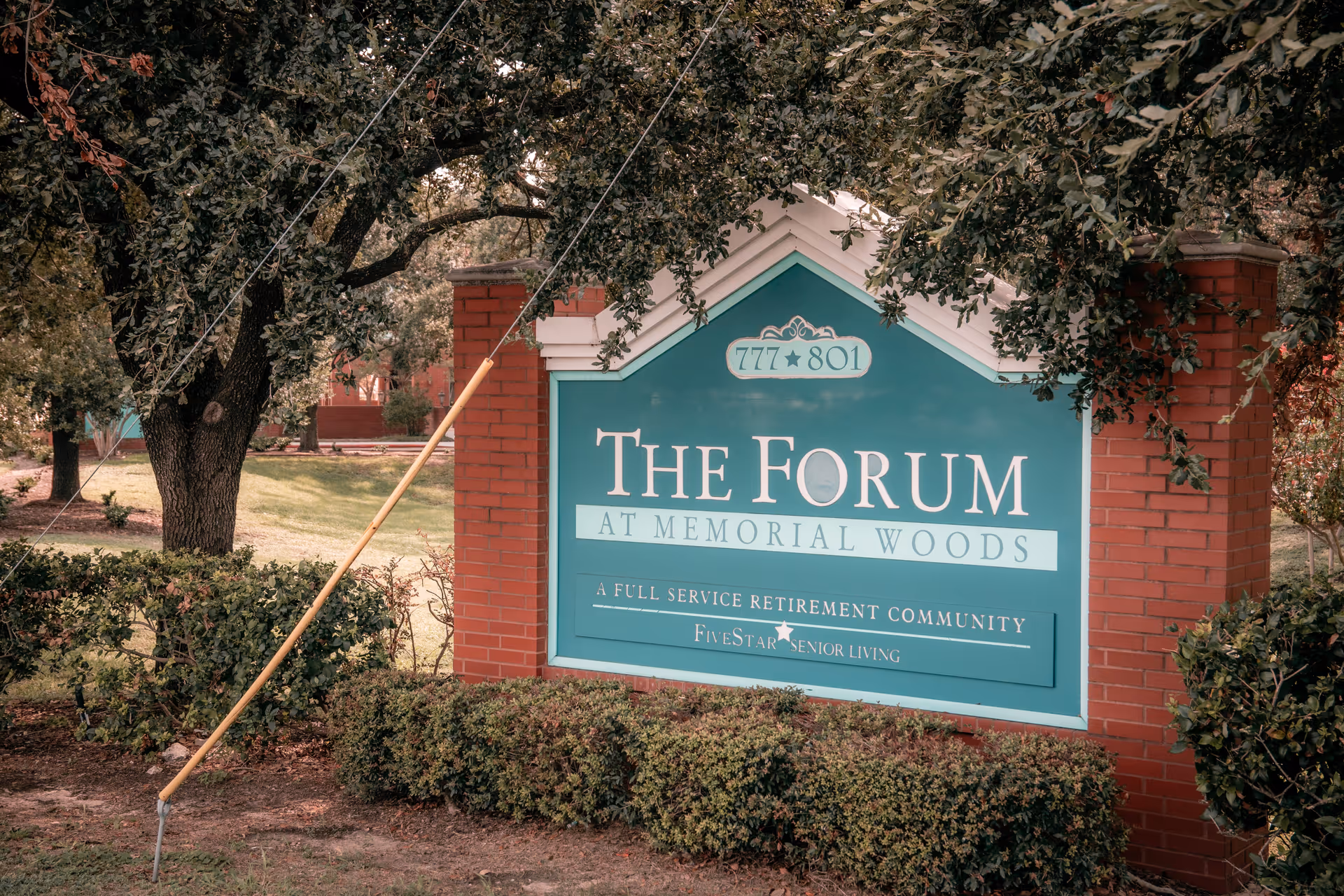 A large green and white sign for The Forum at Memorial Woods, a full service retirement community by Five Star Senior Living, set between two red brick pillars with trees and bushes surrounding it.