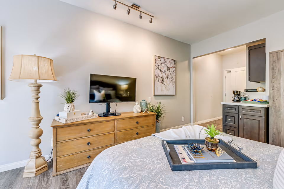 A cozy bedroom in a senior living facility featuring a wooden dresser with a flat-screen TV, decorative plants, and a lamp. The bed has a patterned bedspread with a tray holding books and small plants. The room has light-colored walls, a floral painting, and an open doorway leading to a kitchenette area with dark cabinets and a coffee maker.