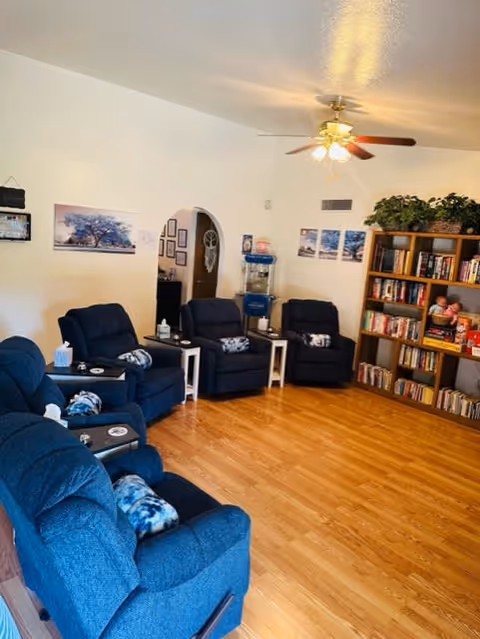 A cozy living room with five blue recliner chairs arranged in a semi-circle on a wooden floor. Each chair has a small side table with tissues and other items. A ceiling fan with lights is on the ceiling. On the right side, there is a tall bookshelf filled with books and some decorative items, including plants on top. The walls are decorated with framed pictures and a water dispenser is visible near an arched doorway.