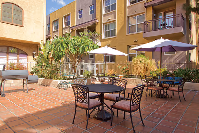 Outdoor patio area with terracotta tile flooring featuring round metal tables with cushioned chairs and umbrellas. There is a barbecue grill on the left side and various plants and small trees around the perimeter. The background shows a multi-story building with balconies and windows under a clear blue sky.