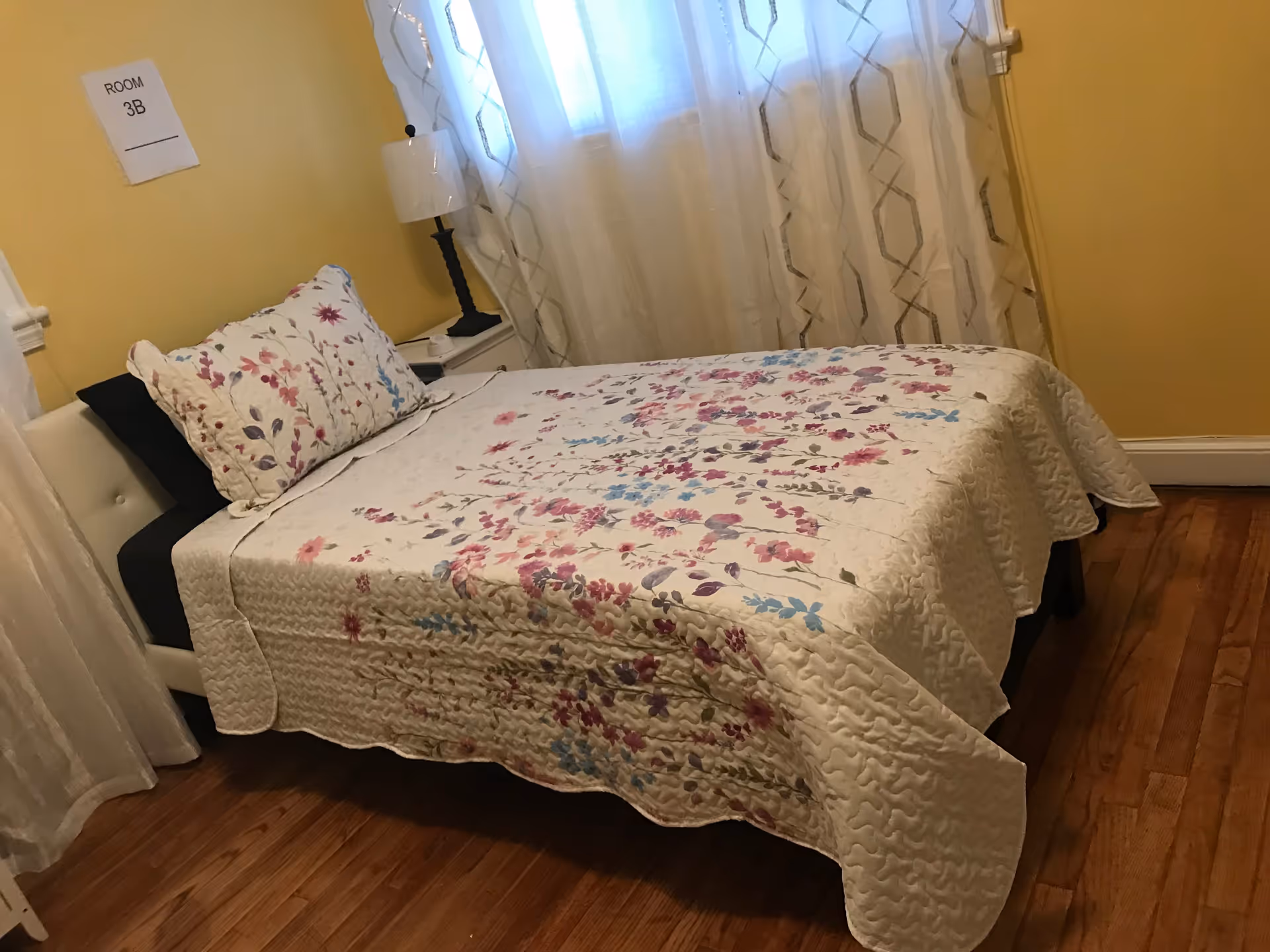 A bedroom with a bed covered in a white quilt with pink, purple, and blue floral patterns. There is a matching floral pillow and a black pillow behind it. A white nightstand with a lamp is next to the bed. The walls are painted yellow and there are sheer white curtains with a geometric pattern on the window. A sign on the wall reads 'ROOM 3B'. The floor is wooden.
