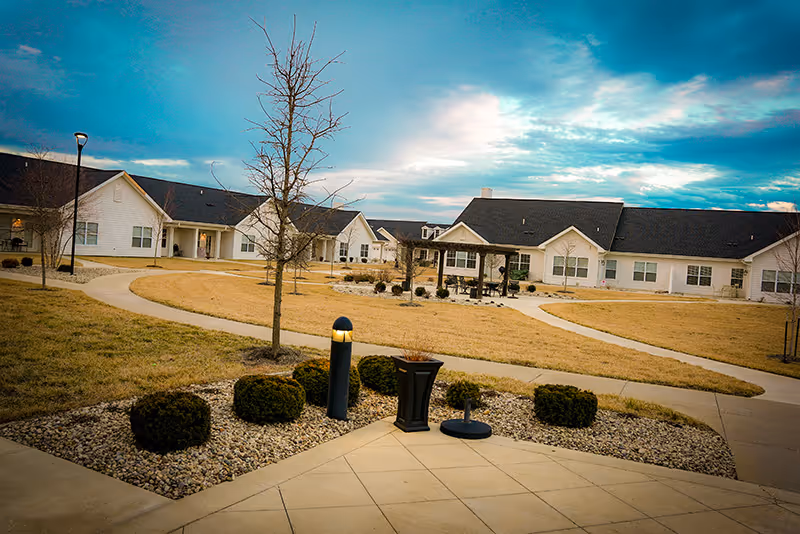 A landscaped courtyard with winding walkways, trimmed shrubs and a gazebo surrounded by single-story white senior living buildings under a blue sky.