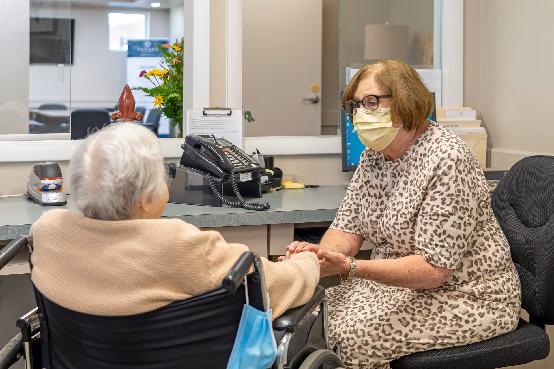 An elderly woman in a wheelchair is holding hands and talking with a healthcare worker wearing a face mask and leopard print dress in an office setting with a desk, telephone, and computer.