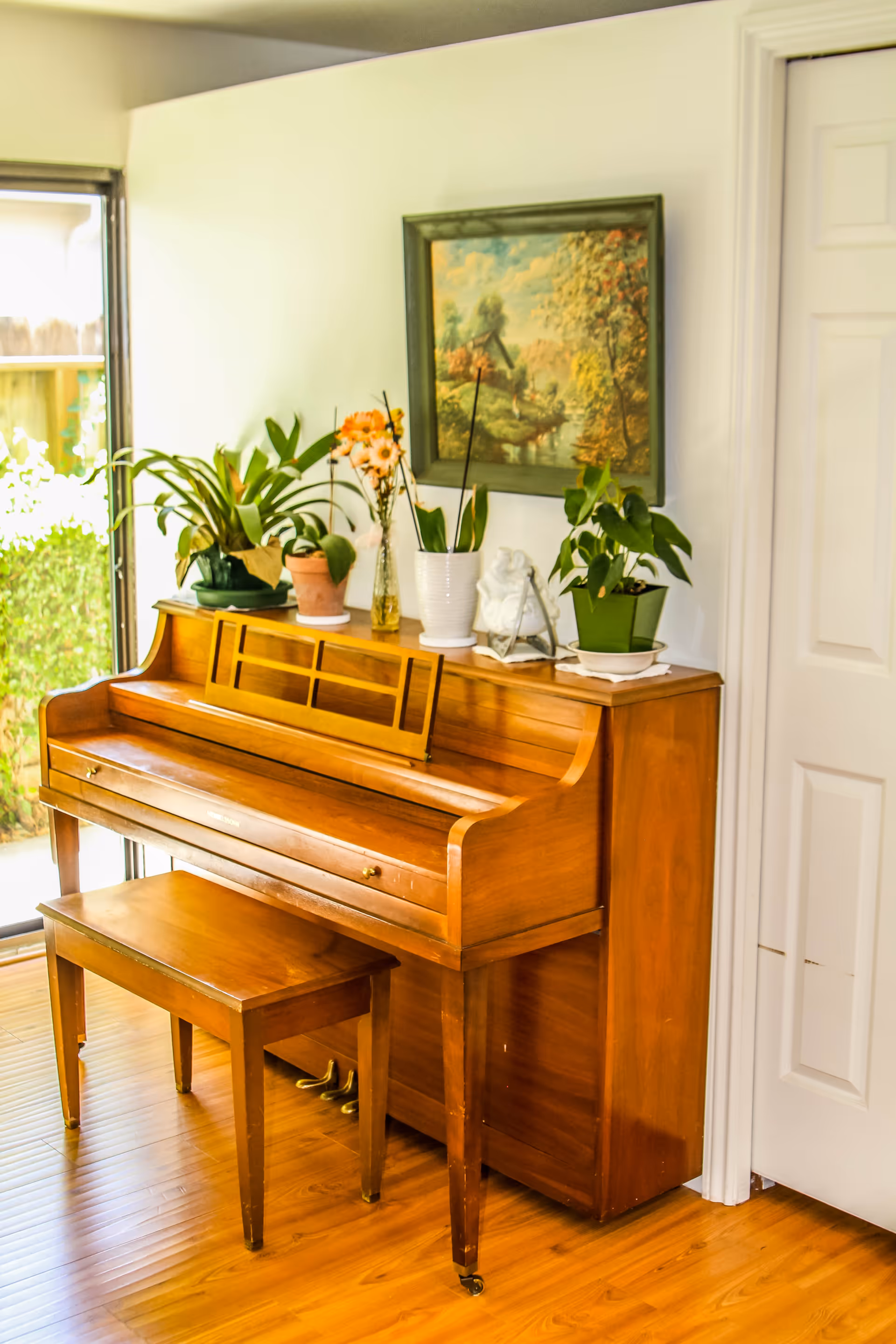 A wooden upright piano with a matching bench in a room with wooden flooring. On top of the piano are several potted plants and a small statue. A framed painting depicting a countryside scene hangs on the wall above the piano. To the left, there is a sliding glass door with a view of greenery outside.