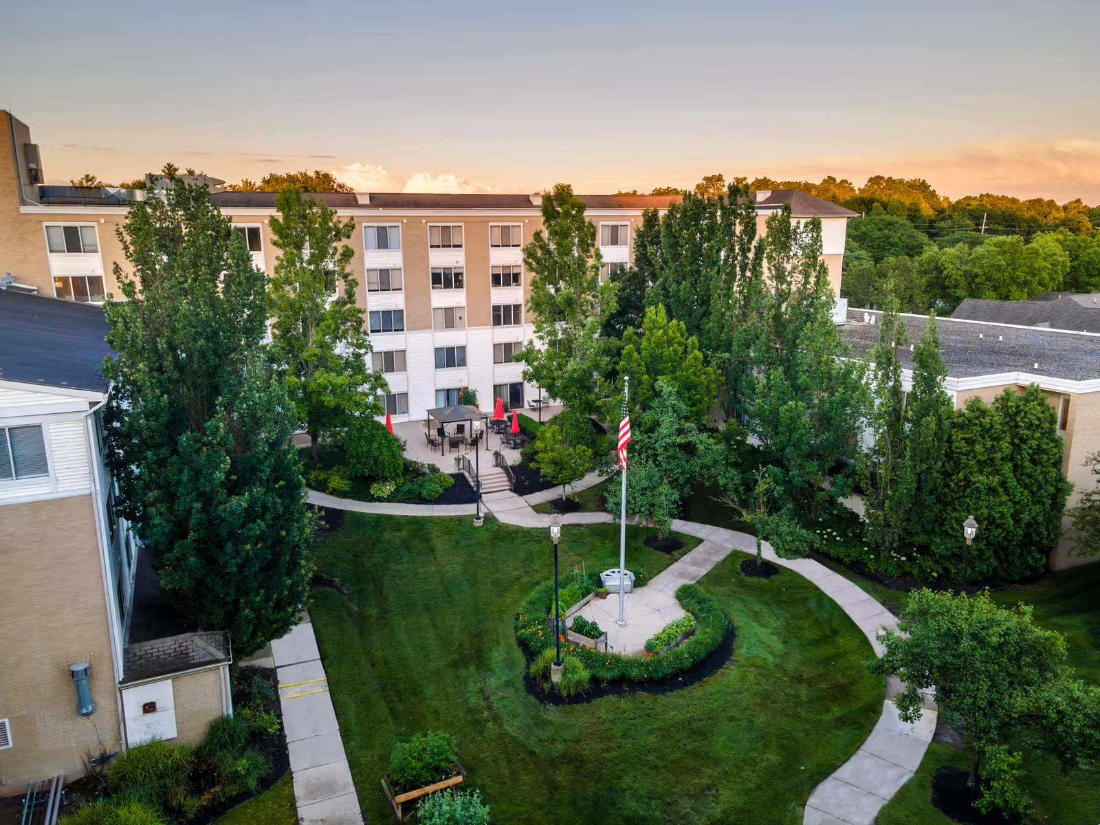 Aerial view of the courtyard at Independence Village of Plymouth, featuring a circular walkway around a flagpole with an American flag, surrounded by well-maintained green lawns, trees, and shrubs. The multi-story residential building with balconies and windows encloses the courtyard, with outdoor seating areas visible near the building.