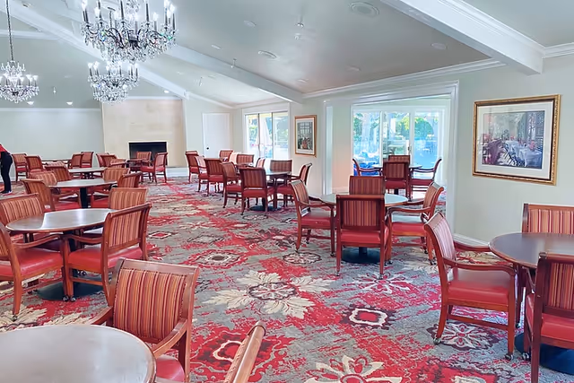 Large dining room with round tables, red upholstered chairs, chandeliers, and a patterned carpet.