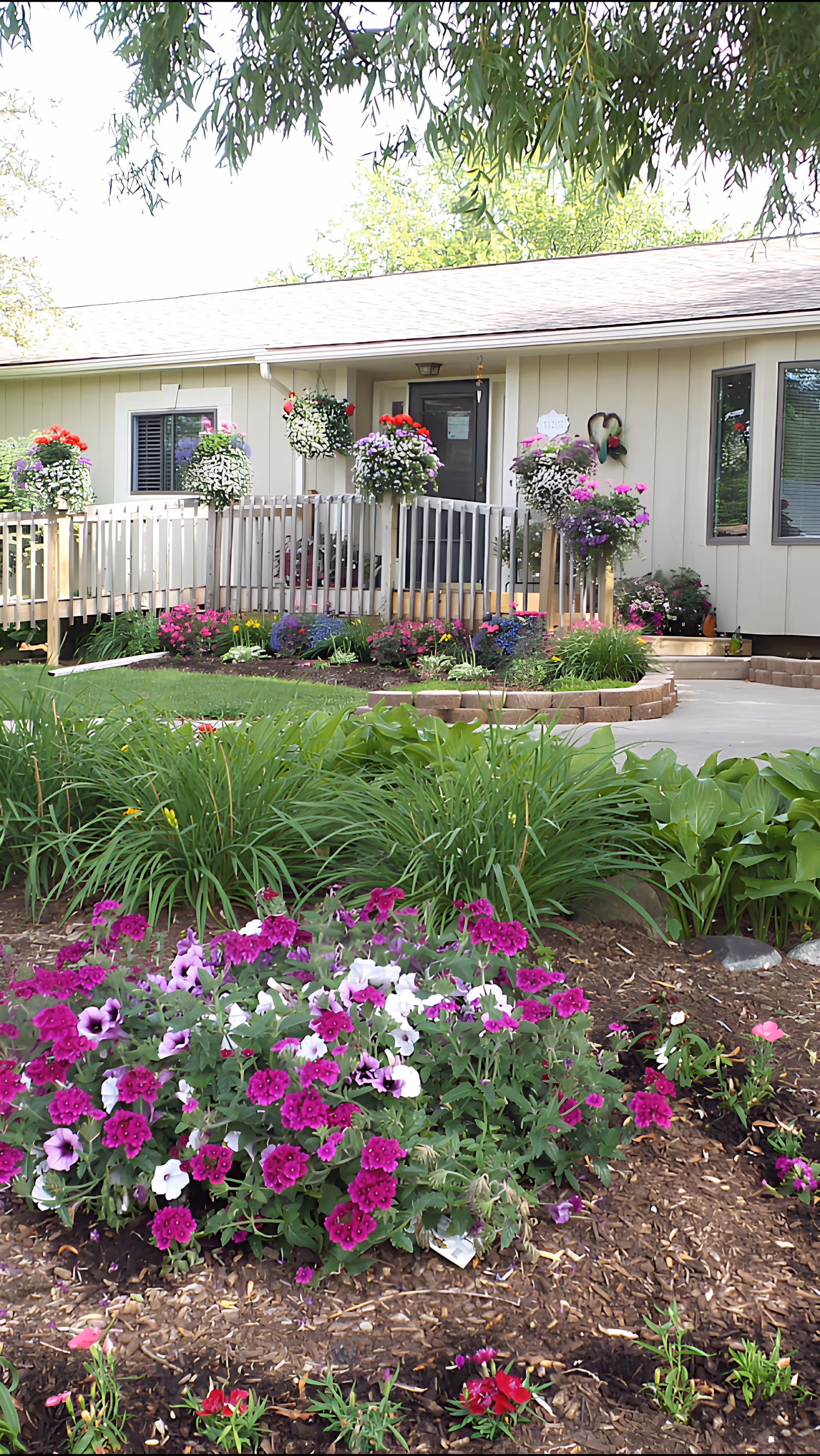 A single-story building with a light beige exterior and a gray door, surrounded by a well-maintained garden featuring vibrant purple, white, and pink flowers. There is a wooden ramp with railings leading to the entrance, and hanging flower baskets adorn the porch area. Green shrubs and trees frame the scene, creating a peaceful outdoor environment.