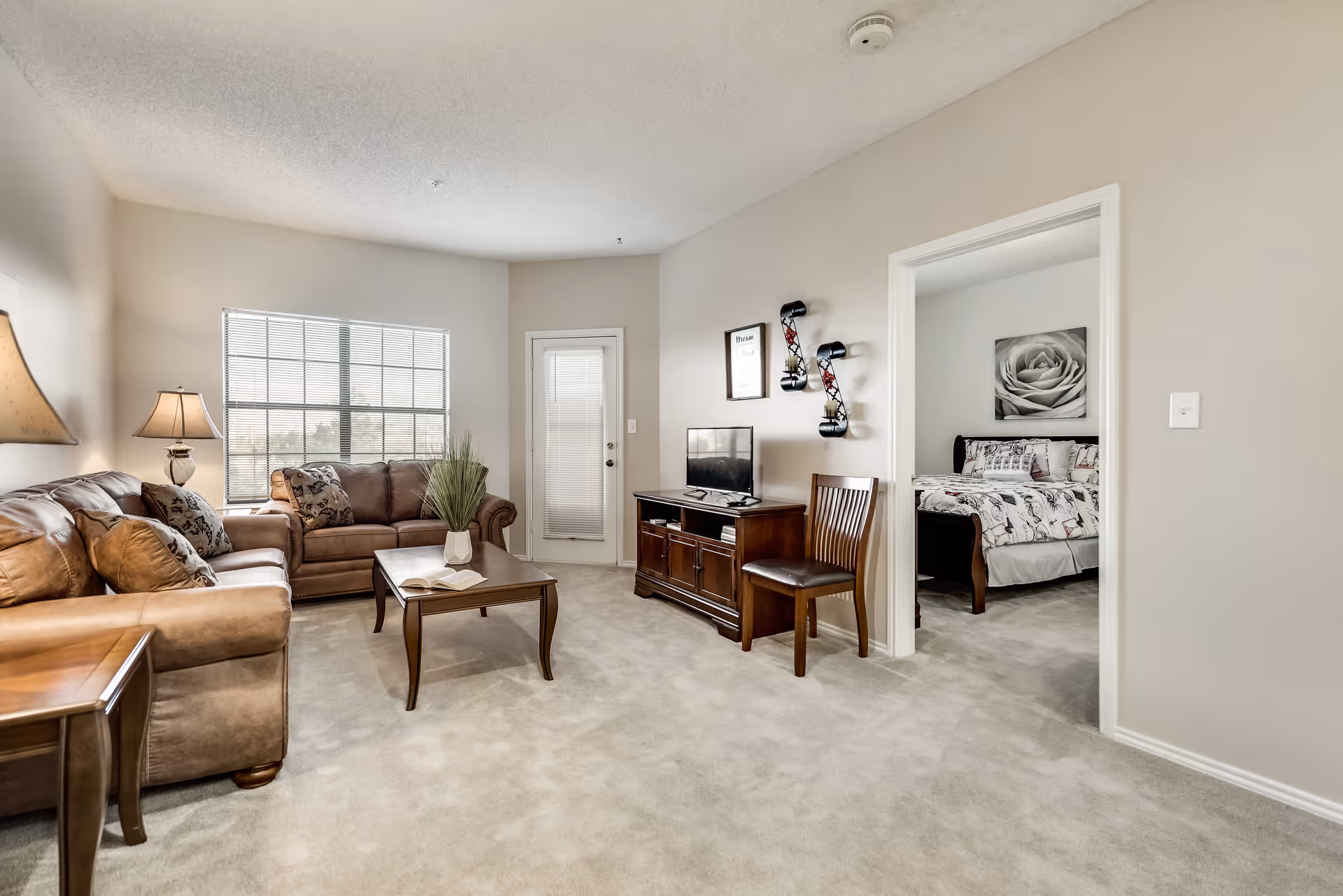 A cozy living room with beige carpet and walls, featuring two brown leather sofas, a wooden coffee table with a book and a vase of greenery, a wooden TV stand with a flat-screen TV, a wooden chair, and a lamp. A large window lets in natural light. An open doorway leads to a bedroom with a bed, floral bedding, and a black and white rose wall art.