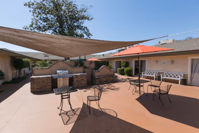 Sunny outdoor courtyard with metal tables and chairs, red umbrellas, a shade sail, and a built-in brick barbecue area.