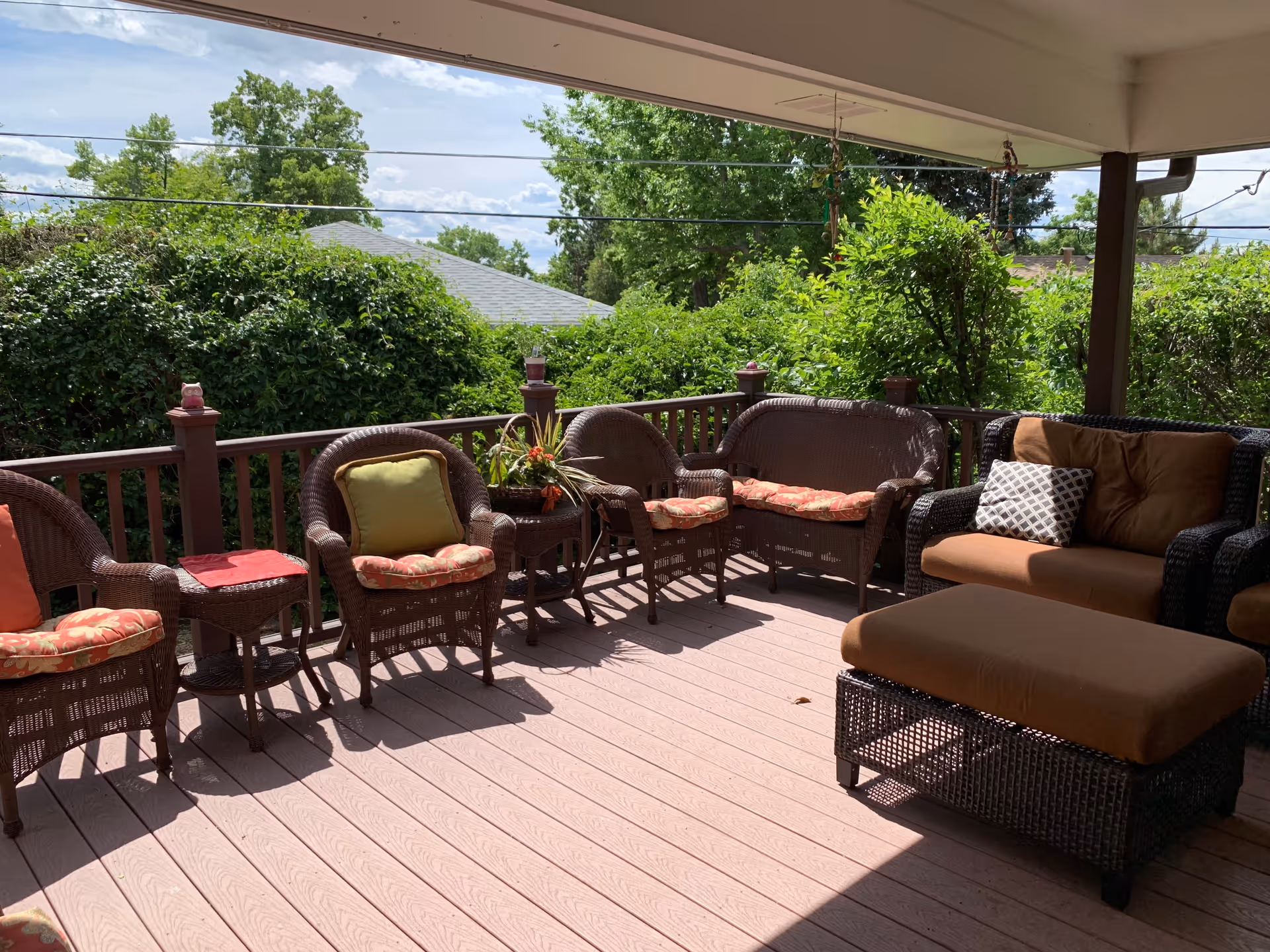 Covered outdoor patio area with wicker furniture including chairs, a loveseat, and an ottoman. Cushions in various colors are on the furniture. The patio overlooks green bushes and trees under a partly cloudy sky.