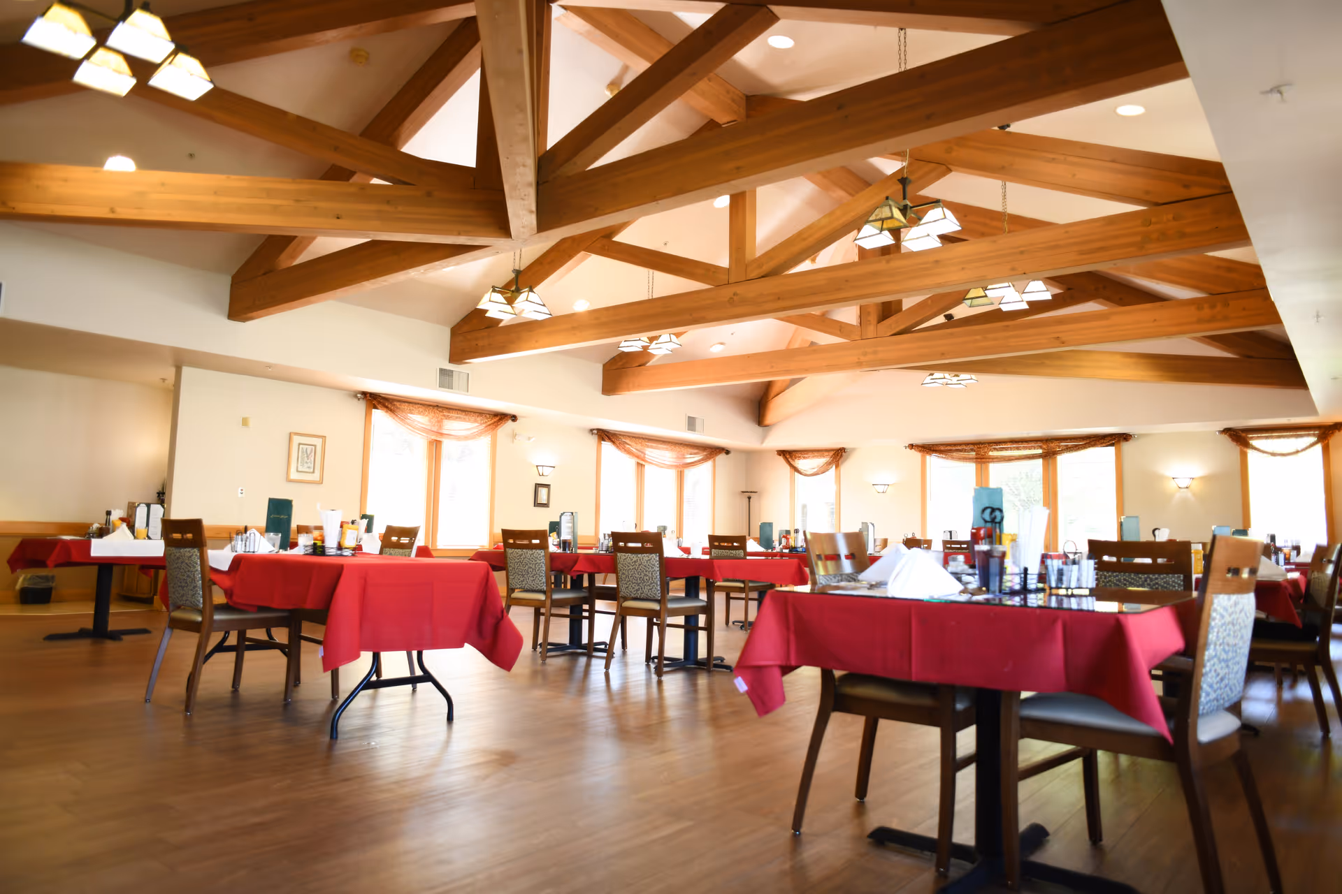 A spacious dining room with wooden beams on the ceiling, multiple tables covered with red tablecloths, and chairs arranged around them. The room has large windows with sheer curtains allowing natural light to fill the space.