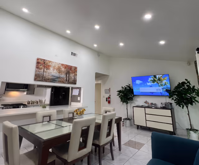 Interior view of a modern assisted living facility dining and living area with a glass-top dining table surrounded by six beige cushioned chairs. A kitchen pass-through window is visible with a black refrigerator and stove. A large painting of autumn trees hangs above the pass-through. On the right side, a flat-screen TV is mounted on the wall above a white and brown cabinet, flanked by two tall green plants. The floor is tiled with white and beige tiles, and the ceiling has recessed lighting.