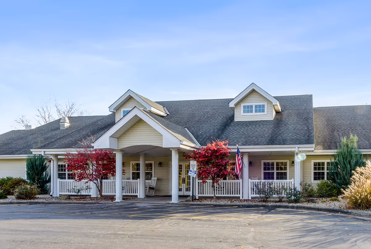 Front exterior view of a single-story building with a covered entrance supported by white columns. The building has a gray shingled roof with dormer windows and beige siding. There are two small red-leafed trees and some shrubs in front of the building, along with an American flag near the entrance. The sky is clear and blue.