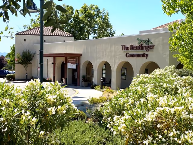 Exterior view of The Reutlinger Community building with beige walls, arched entryways, and a red-tiled roof, surrounded by green trees and bushes with white flowers under a clear blue sky.