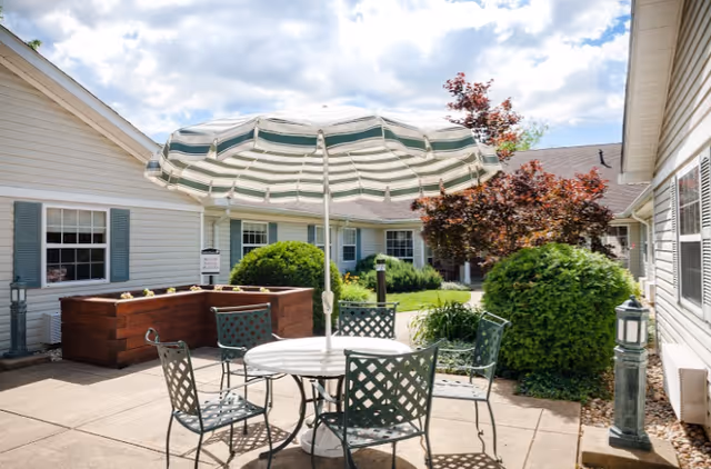 Outdoor patio area with a round white table and four metal chairs with lattice backs. A large green and white striped umbrella shades the table. Surrounding the patio are beige buildings with blue shutters, green bushes, and a small tree with red leaves under a partly cloudy sky.