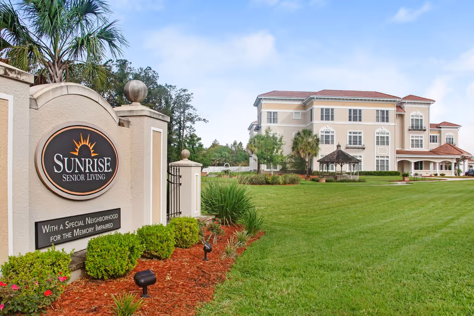 Exterior view of Sunrise Senior Living facility in Jacksonville showing a large beige building with multiple windows and a red roof, a well-maintained green lawn, a gazebo, and a sign at the entrance that reads 'Sunrise Senior Living with a special neighborhood for the memory impaired'.