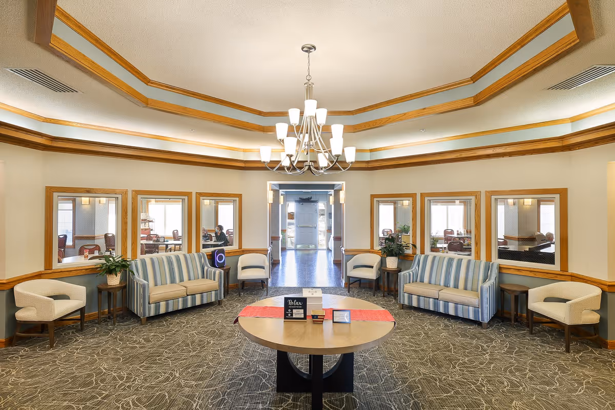 A spacious and well-lit common area in an assisted living facility with two striped sofas, four beige armchairs, a round wooden table with a red runner and informational signs, and a large chandelier hanging from a tray ceiling with wooden trim. Several windows on the walls provide views into adjacent rooms with tables and chairs.