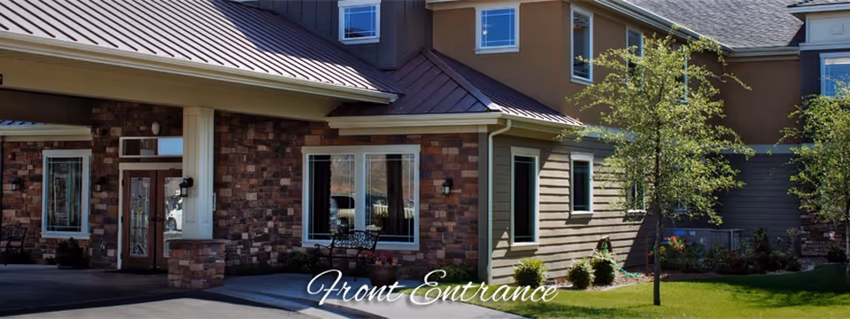 Front entrance of a senior living building with stone and siding facade, covered entryway, windows, bench and landscaped lawn.