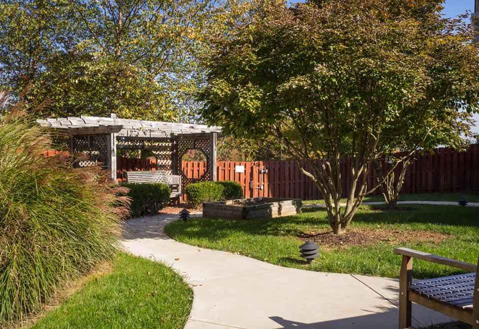 A sunny outdoor garden area with a curved concrete pathway leading to a wooden pergola with a bench underneath. The garden is surrounded by green grass, bushes, and trees, with a wooden fence in the background.
