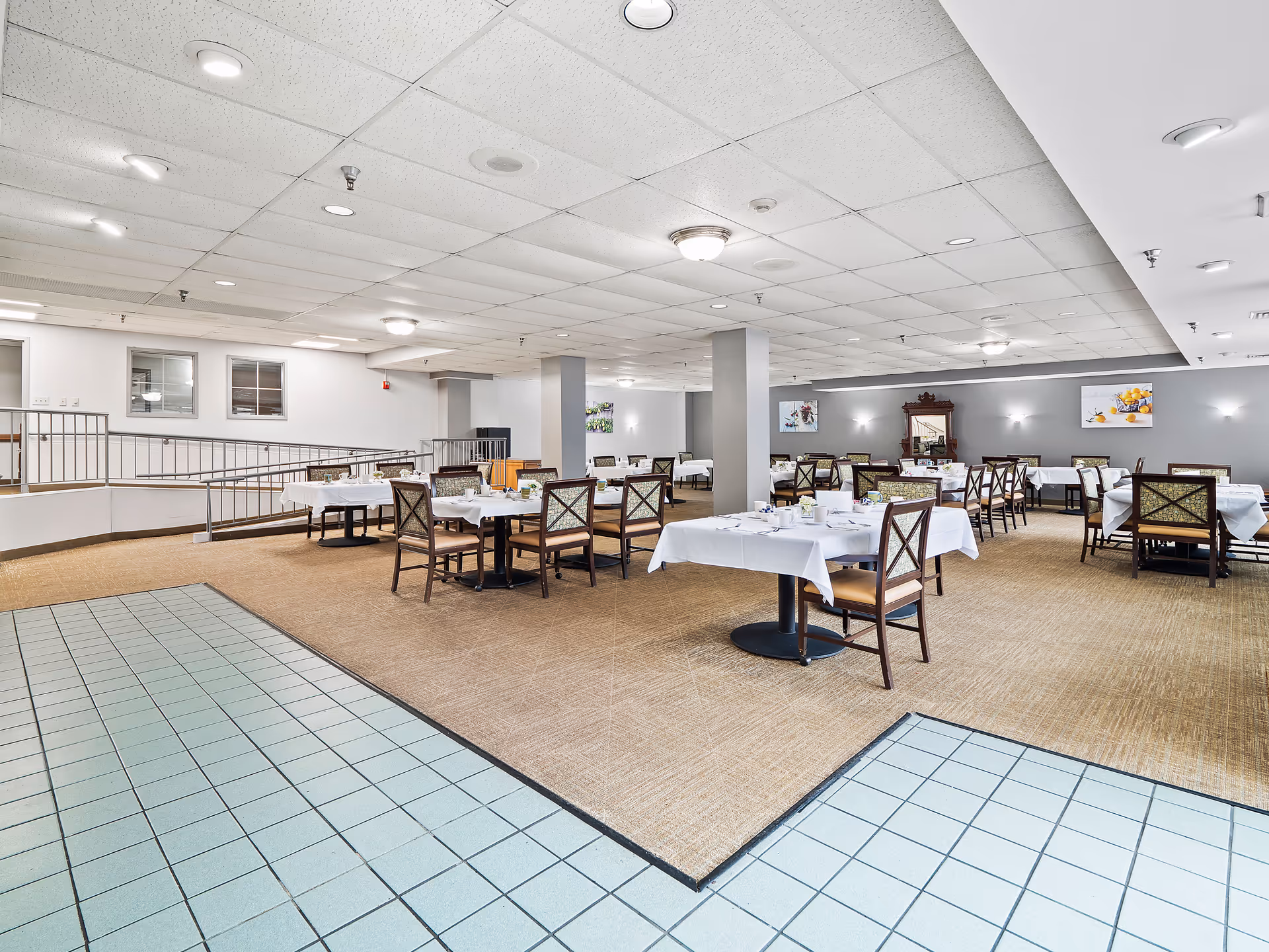 A spacious dining room with multiple tables covered in white tablecloths, each set with cups, plates, and small flower arrangements. The room has beige carpet flooring with a tiled area in the foreground, gray walls with framed artwork, and a ceiling with recessed lighting. There are wooden chairs with patterned upholstery around the tables.