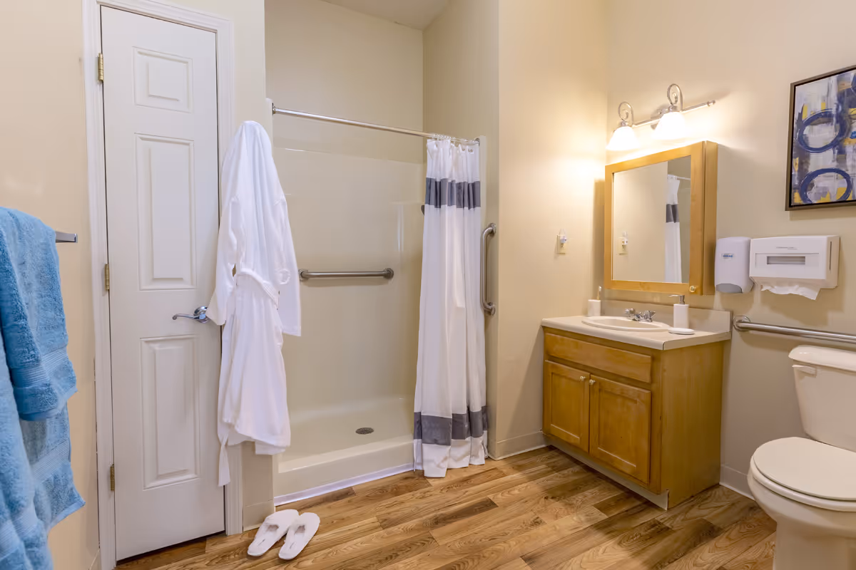 A clean bathroom with a white shower curtain, a white bathrobe hanging on the door, blue towels on a rack, a wooden vanity with a sink and mirror, a toilet, and hardwood flooring.
