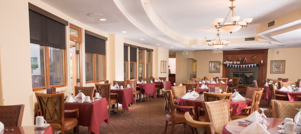 Spacious dining room with round and rectangular tables covered in burgundy tablecloths, place settings and chairs, large windows, and ceiling chandeliers.